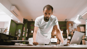 Man standing at a desk, reviewing printed documents with a laptop and office supplies around him, representing comparison shopping and decision-making.