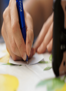 Hand holding a blue pen writing on a small piece of paper.