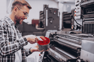 Man working with a printing press, holding a container of red ink and preparing it for use in the machine