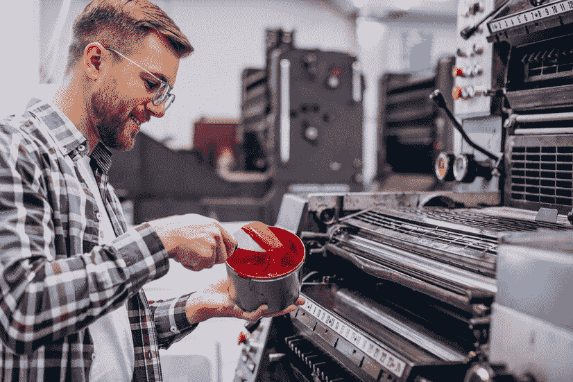 Man working with a printing press, holding a container of red ink and preparing it for use in the machine