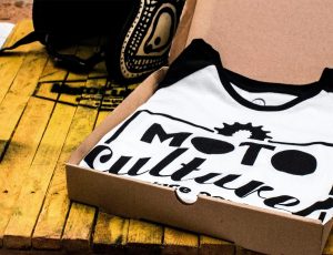 A white and black ringer T-shirt with a "MOTO culture" logo inside a cardboard shipping box.