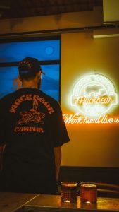 Man from behind in a Mackbear Company shirt looking at a neon Mackbear Coffee sign.