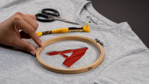 Hand adjusting a wooden embroidery hoop over a red appliqué letter on a gray shirt.