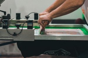 Hands using a squeegee to push ink across a screen-printing mesh frame.