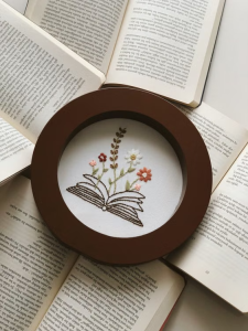Round wooden hoop framing an embroidered open book with colorful flowers sprouting from its pages.